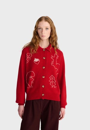 Jeune femme aux longs cheveux roux ondulés porte un cardigan rouge boutonné avec des motifs blancs d'animaux et de feuilles et un pantalon sombre, debout devant un fond uni.
