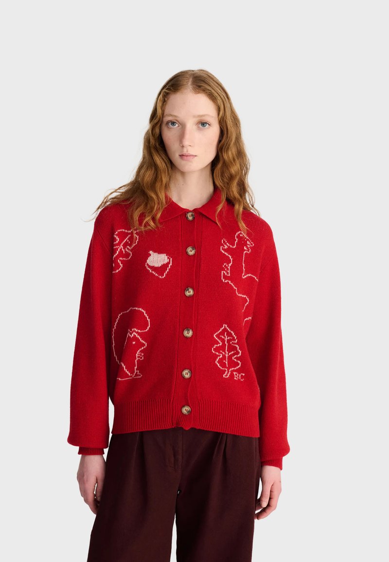 Jeune femme aux longs cheveux roux ondulés porte un cardigan rouge boutonné avec des motifs blancs d'animaux et de feuilles et un pantalon sombre, debout devant un fond uni.