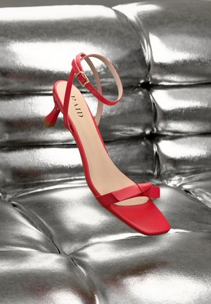 Sandale rouge à talon avec un bout carré, une fine bride de cheville et un détail de nœud. Fabriquée dans un matériau lisse, elle présente une boucle dorée et un design élégant.