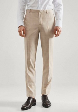 Mand iført beige slim-fit dressbukser med svagt ternet mønster, hvid skjorte med lange ærmer og sorte lædersko står.