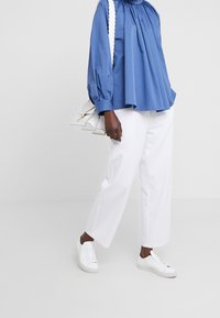 Personne à la peau foncée portant un chemisier bleu à manches longues, un pantalon blanc court, des baskets blanches et tenant un sac à main blanc sur un fond blanc.