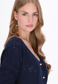 Cardigan tricotat de culoare albastru marin, cu decolteu în V, cu un model texturat și trei nasturi din sidef în față. Material moale și ușor.