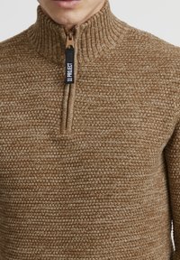 Maglione lavorato a maglia marrone con colletto alto, dotato di mezza zip e una texture intrecciata. Tirazip nero con etichetta bianca.