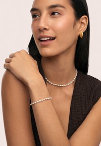 Parelketting en armband sieren een model. De halslijn is versierd met kleine, ronde parels; de armband is een match. Oorbellen in de vorm van goudbladeren completeren de look.