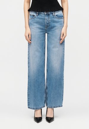 Femme portant un jean bleu clair à jambes larges et des talons hauts noirs pointus, debout devant un fond blanc uni.