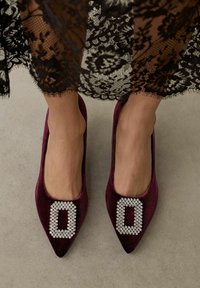 Pies con zapatos planos puntiagudos de terciopelo burdeos con hebillas rectangulares de pedrería, de pie sobre un suelo gris claro bajo el dobladillo de un vestido negro de encaje.