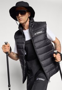 Black puffy vest with high collar, logo, and zipper. Worn over a white shirt, paired with pants and a black bucket hat and sunglasses.