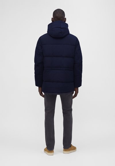 Un homme portant une doudoune bleu marine à capuche, un pantalon gris foncé et des chaussures couleur beige, debout, de dos, sur un fond blanc uni.
