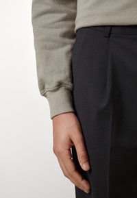 Šedá mikina s žebrovanými manžetami, spárovaná s tmavými kalhotami. Detailní záběr ukazuje texturu látky a ruku spočívající na nohavici kalhot.