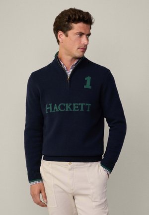 Donkerblauwe gebreide trui met een kwart rits, voorzien van een groene "1" en het "HACKETT" logo. Zachte textuur, geribbelde manchetten en zoom.