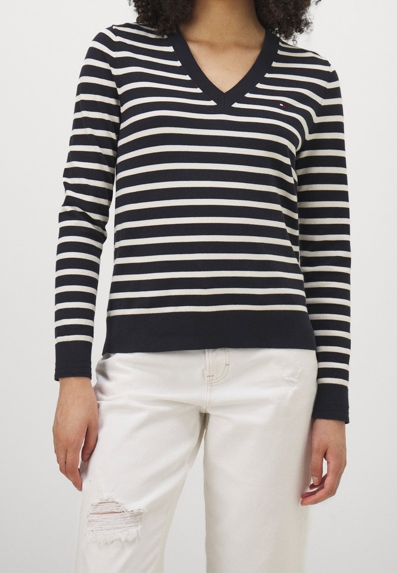 Personne portant un haut à manches longues rayé noir et blanc avec un col en V et un jean blanc déchiré sur un fond uni.