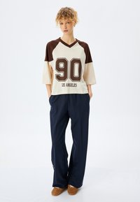 Top oversized en crema y marrón con el número "90" y la impresión "LOS ÁNGELES", combinado con pantalones de pierna ancha en azul marino y zapatos tipo slip-on marrones.