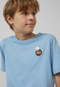 Lichtblauwe katoenen t-shirt met een ronde zwarte patch met een lachend gezicht en de tekst "SMILEY WORLD", geplaatst op de linkerborst.