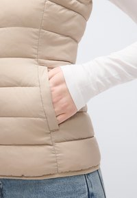 Μπεζ αμάνικο μπουφάν με καπιτονέ υφή και πλαϊνή τσέπη. Φορεμένο πάνω από λευκό μπλουζάκι με μακριά μανίκια, μέσα σε μπλε τζιν.