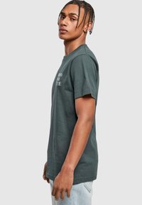 Mørkegrøn bomulds T-shirt med korte ærmer, rund halsudskæring og hvid tekstgrafik foran, fremvist på en model i profil.