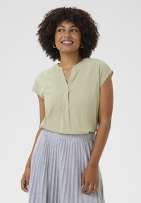 Blusa verde chiaro a maniche corte con colletto alla mandarino e piega centrale. Abbinata a una gonna grigia plissettata con un leggero motivo a righe.