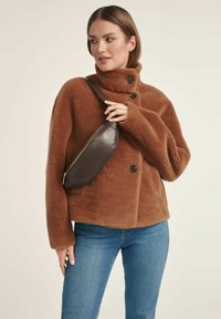 Kobieta ubrana w brązową, puszystą kurtkę i niebieskie dżinsy, trzymająca ciemnobrązową skórzaną torbę na ramię na piersi.