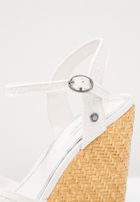 Sandale blanche à talon compensé en paille tissée, bride de cheville avec boucle argentée et petit détail métallique rond marqué sur la bride.