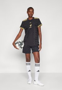 Svart fotballdrakt med gullaccentuering, med et teksturert mønster og en logo. Båret med svarte shorts, hvite sokker og fotballsko, mens han holder en ball.