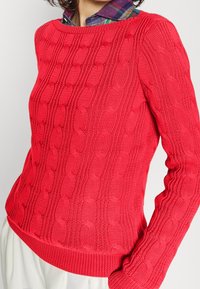 Röd stickad tröja med strukturerat flätmönster, rund halsringning och ribbade muddar och fåll. Långa ärmar med en figurnära design.