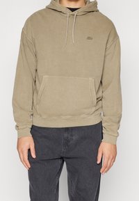 Felpa beige realizzata in tessuto morbido, con tasca a marsupio, cappuccio regolabile e un piccolo logo sul petto. Vestibilità comoda con polsini a coste.