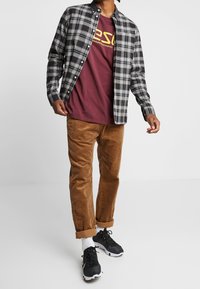 Koszula w kratę w czarno-szarym kolorze noszona na bordowej koszulce z grafiką. Beżowe spodnie z korduroyu o teksturowanym wykończeniu oraz czarne sneakersy z białymi akcentami.