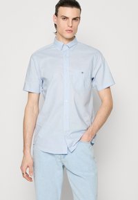 Homme portant une chemise bleu clair à manches courtes boutonnée et un jean bleu clair, debout avec une main dans la poche, sur un fond uni.