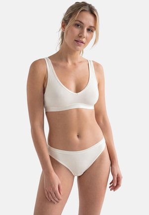 Kvinde iført en cremefarvet sømløs bh og matchende trusser, stående mod en ensfarvet hvid baggrund med et neutralt udtryk.