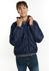 Giacca puffer blu navy con una texture liscia e lucida, caratterizzata da una chiusura con zip e polsini elastici. Indossata sopra un maglione a fantasia e jeans di denim.