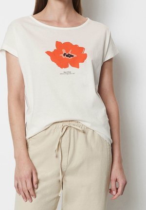 Kvinde iført hvid T-shirt med stort rød-orange blomsterprint og tekst, kombineret med beige bukser med snøre, vist fra skuldre til hofter.