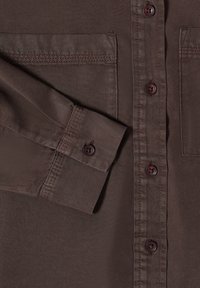 Chemise marron à manches longues avec poignets boutonnés, boutons à l'avant et poches poitrine, montrant des coutures détaillées et la texture du tissu.