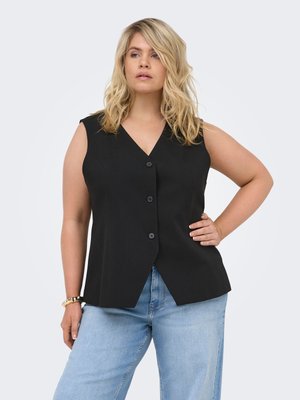Blond kvinne iført en ermeløs svart bluse med knapper og lyseblå jeans, poserer med en hånd på hoften mot en ensfarget bakgrunn.
