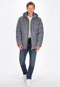 Jachetă puffer gri cu glugă, design matlasat, închidere cu fermoar în față și buzunare. Purtați peste o cămașă albă, asortată cu blugi întunecati și adidași albi.