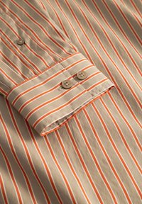 Chemise à rayures beige avec de fines lignes orange, dotée d'un poignet à boutons et d'une texture de tissu douce. Deux boutons visibles sur la zone du poignet.