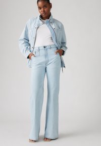 Világos kék farmerdzseki magas gallérral, hozzá illő, magas derékú, bólogató farmernadrággal párosítva. Simító textúra és minimalista részletezés.