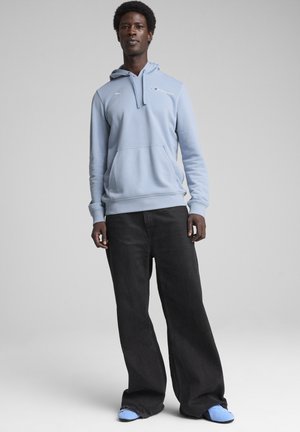 Sudadera con capucha de color azul claro, con un bolsillo delantero, puños de canalé y un pequeño logotipo en el pecho, combinada con jeans negros de pierna ancha y zapatillas azules.