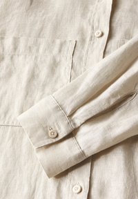 Camicia in lino beige chiaro con colletto e polsini abbottonati, caratterizzata da una superficie testurizzata e due tasche sulla parte anteriore.