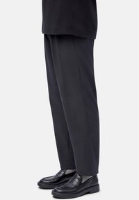 Person wearing dark gray straight-leg pants with black leather loafers and light gray socks, standing against a white background.