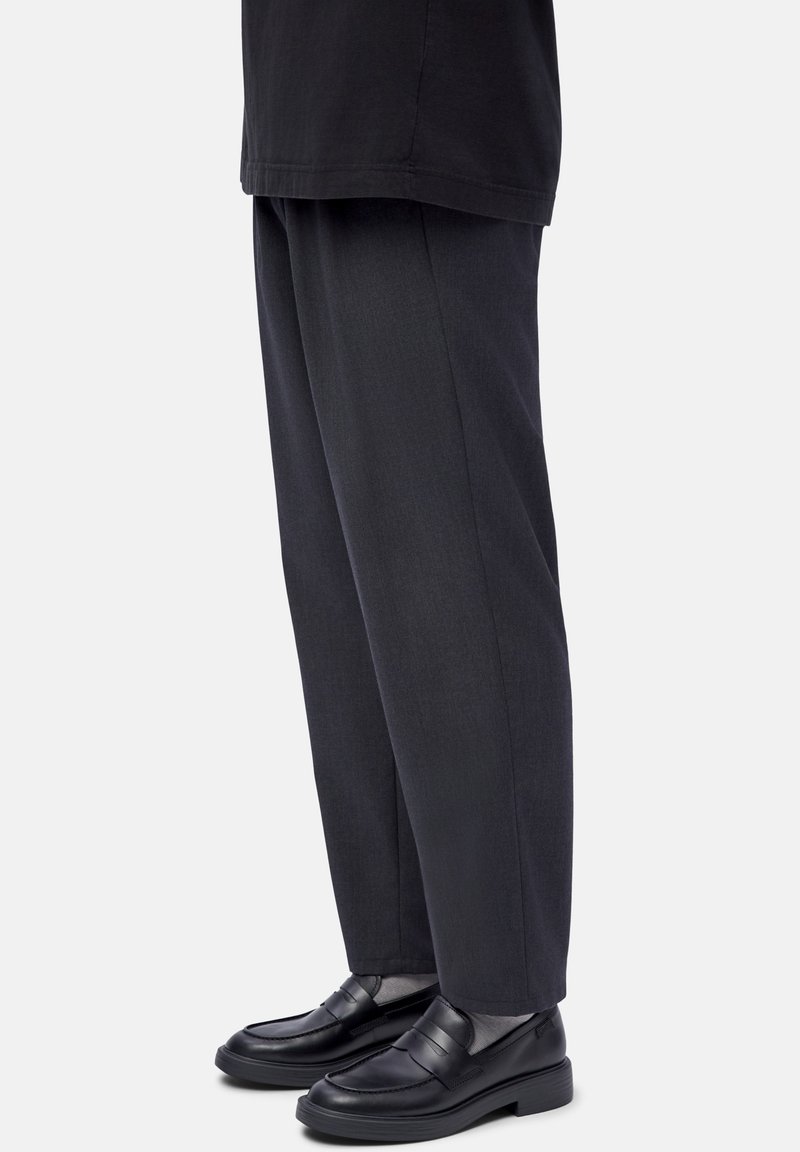 Person wearing dark gray straight-leg pants with black leather loafers and light gray socks, standing against a white background.