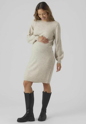 Knit maternity dress in light beige with long, puffed sleeves and ribbed cuffs, paired with knee-high black leather boots.