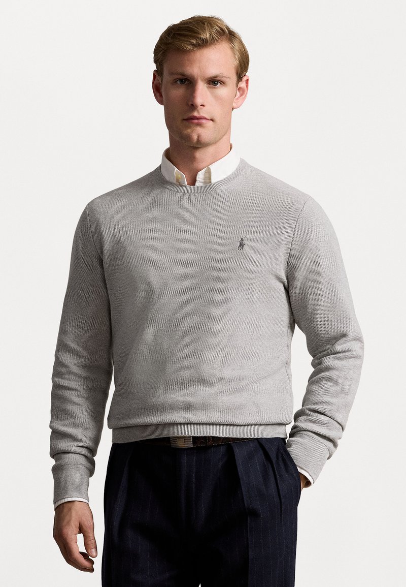Pull en tricot gris clair avec manches longues, col rond et poignets côtelés. Présente un petit logo brodé sur la poitrine. Stylé avec une chemise blanche à col en dessous.