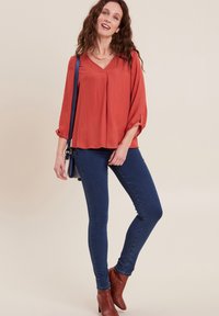 Blusa color ruggine con scollatura a V e maniche a tre quarti, abbinata a jeans aderenti blu scuro e stivaletti marroni alla caviglia. Collana semplice a catena.
