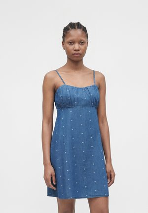 Mujer con el pelo trenzado que lleva un vestido azul sin mangas con tirantes finos y pequeños estampados florales blancos, de pie frente a un fondo liso.