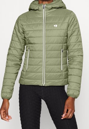 Donna con giacca trapuntata verde chiaro con cerniere bianche e cappuccio, abbinata a leggings neri texturizzati.