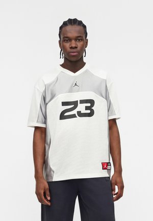 Mladý muž s vrkočmi, oblečený v bielom a sivom športovom drese s číslom 23 a čiernych nohaviciach, stojaci pred bielym pozadím.