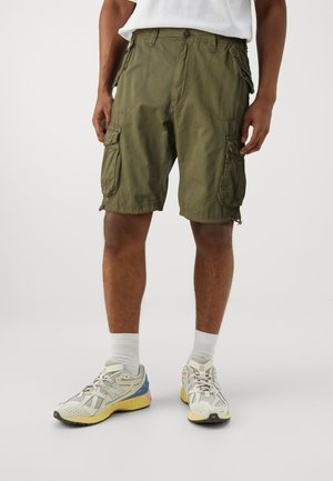 Un homme portant un short cargo vert olive, des chaussettes blanches et des baskets New Balance beiges, jaunes et bleues se tenant devant un fond uni.