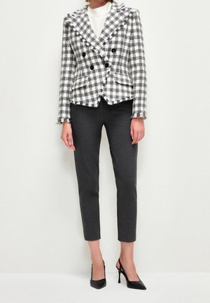 Femme portant un blazer croisé à carreaux noir et blanc, pantalon slim foncé, haut blanc et escarpins noirs à bouts pointus, debout.