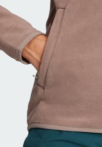 Hellbraune Fleecejacke mit weicher Textur, herausragender Tasche und kontrastierendem Taillenbund. Das Design umfasst einen runden Ausschnitt und enganliegende Bündchen.
