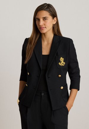 Kvinde iført sort dobbeltradet blazer med guldknapper og broderet våbenskjold, sort top, sorte bukser, hænder i lommerne, neutral baggrund.