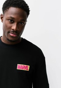 Mustast puuvillast T-särk, millel on roosa ja kollasega ristkülikukujuline Marveli logo. Lihtne ümara kaelusega ja pehme kangaga.